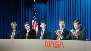 five men sit at a long desk and are all looking at the man on the right who is holding up a space shuttle model for a demonstration.