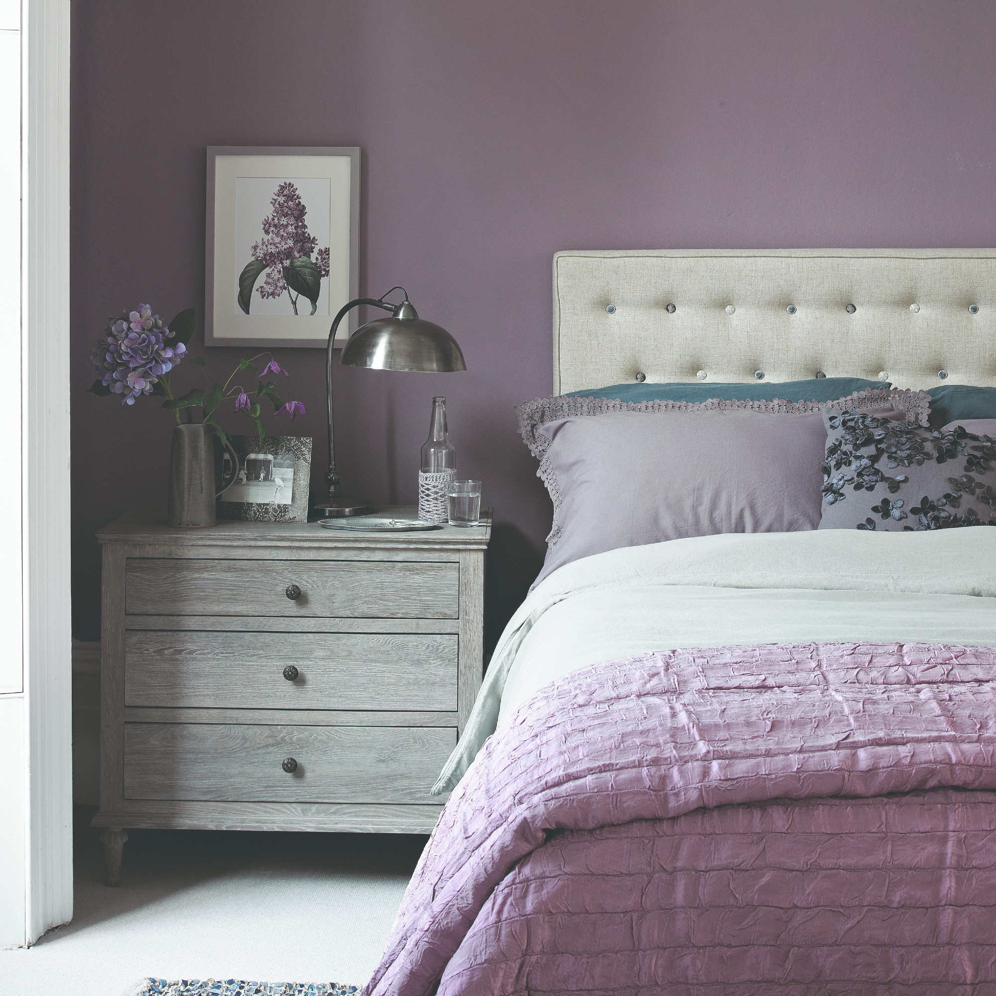 A bedroom painted in lilac with an upholstered off-white bedroom and tonal lilac pillows and bedspread on the bed