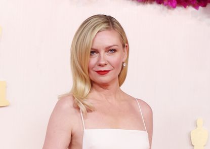 A headshot of Kirsten Dunst on a red carpet wearing a white dress and smiling at the camera
