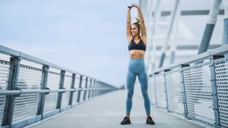 Woman standing stretching overhead while walking across a bridge