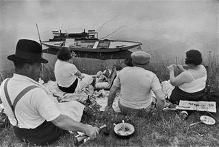 Black and white 1938 French image of two men and two women sat on blankets on a grass verge enjoying a picnic, drinking wine and eating food, to the bottom of the shot is water with a narrow boat floating near to the waters edge