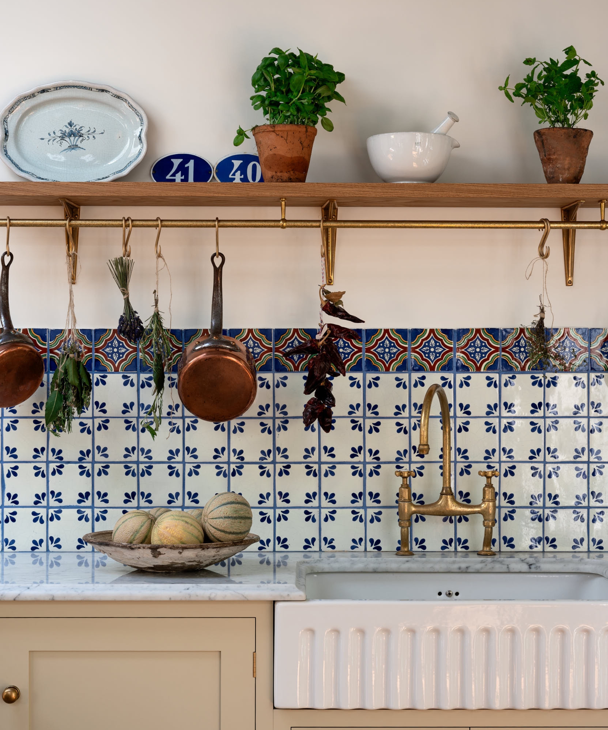 A cream kitchen with a blue and white patterned backsplash and a wooden shelf fit with a brass hanging rail for pots and utensils