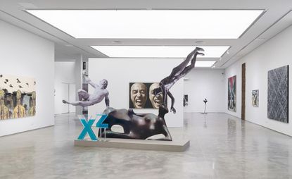 Art exhibition space with white walls and spaced out artwork displayed on the walls. In the foreground of the picture is a large scuplture featuring 3 naked people in a mix of styles.