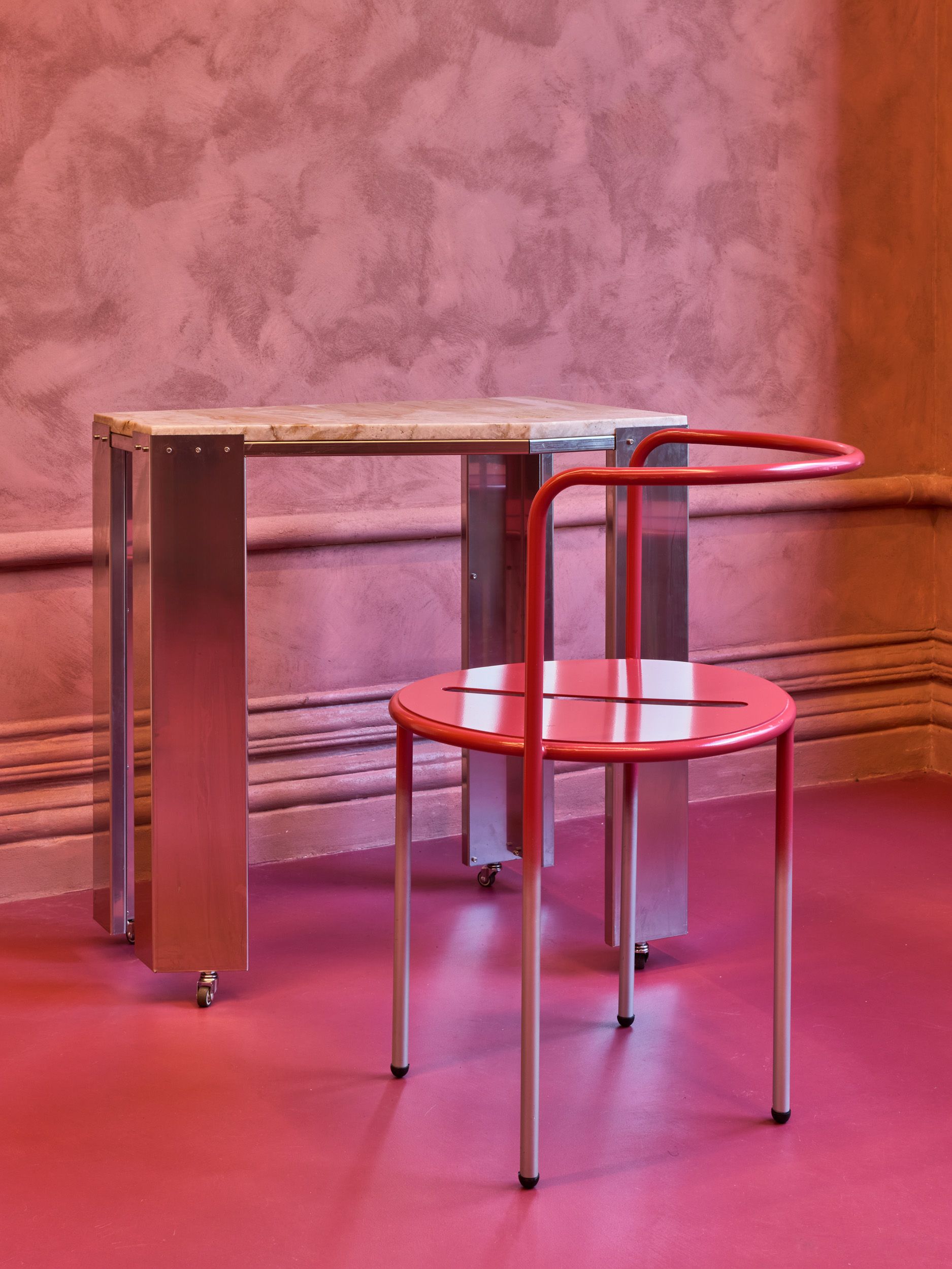 A pink-painted cafe with bright pink and chrome chairs, marble-topped sculptural tables with chrome legs, and lots of deli products and artworks styled on its shelves.