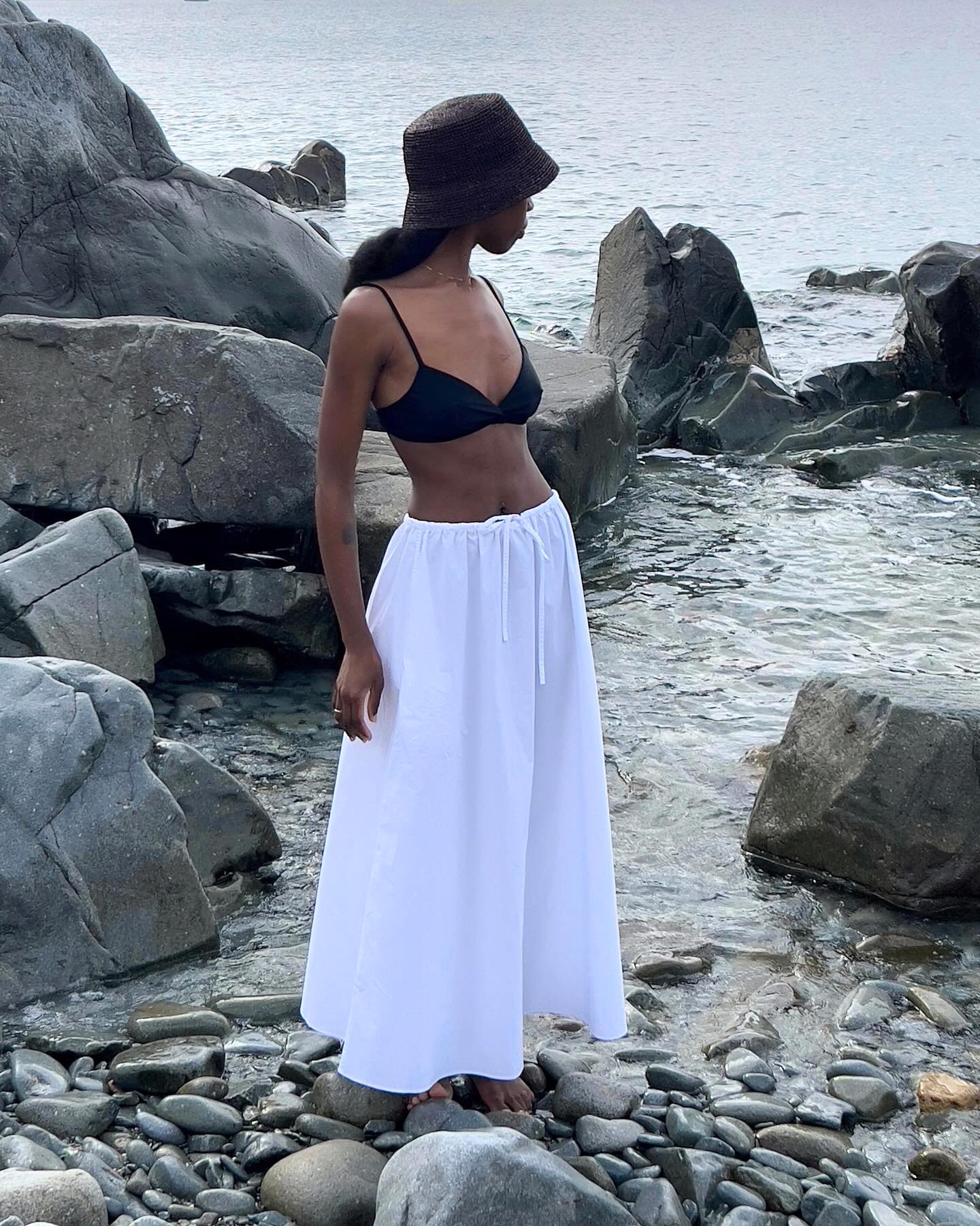 Style influencer Sylvie Mus poses on rocks on a beach in the South of France wearing a raffia black bucket hat, black bikini top, and full white maxi skirt.