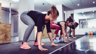 Line of women in gym perform sprawl exercise, bent over with hands on floor, about to jump their feet behind them