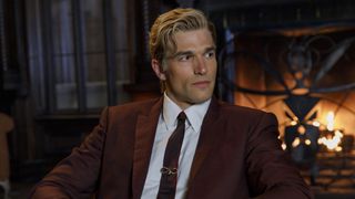 Paul Anthony Kelly's mystery character wearing reddish-brown jacket and tie with a snake clip, sitting in front of a lit-up fireplace inside the Apollyon hotel in American Horror Story 13