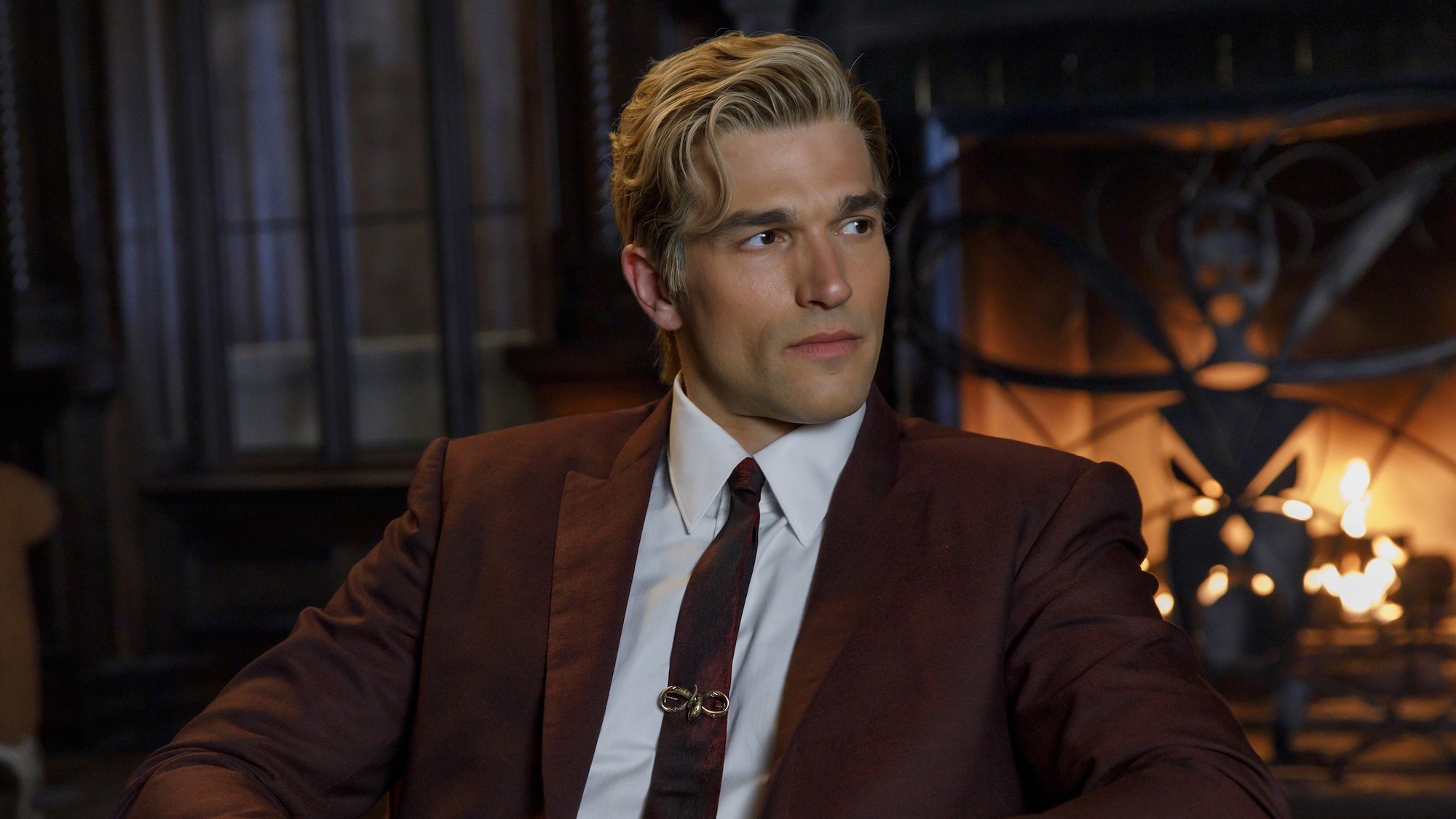 Paul Anthony Kelly's mystery character wearing reddish-brown jacket and tie with a snake clip, sitting in front of a lit-up fireplace inside the Apollyon hotel in American Horror Story 13