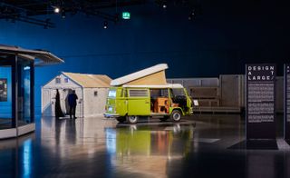 View of the 'Design at Large' exhibition featuring a green 1960s Volkswagen Campervan. There is also a white structure with a pitched roof, a structure with glass windows and a flat roof and black rectangular signs with white text in a space with dark walls and black floors