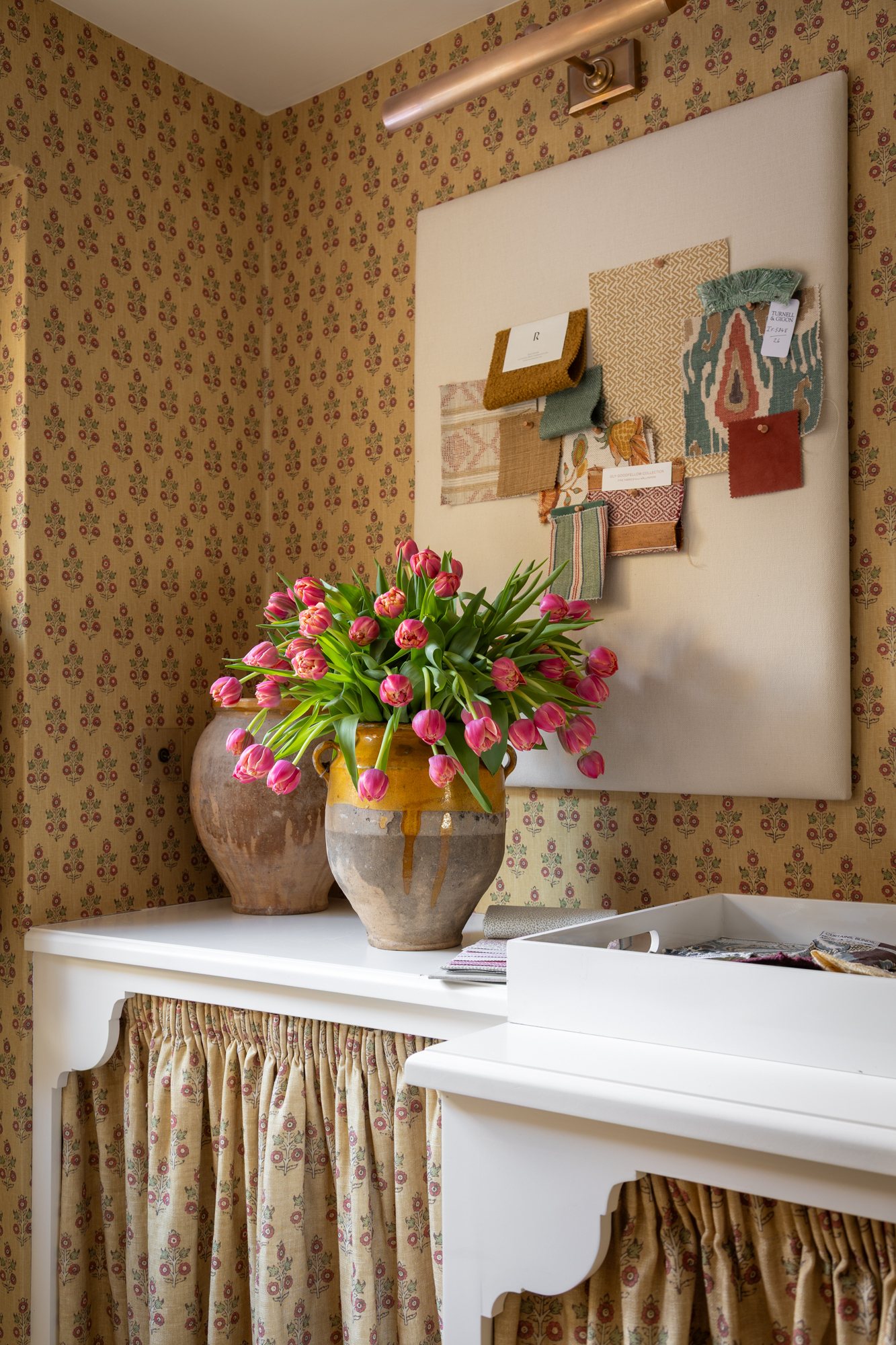A sideboard with a wase of pink tulips, the wallpaper behind is in a ditsy floral print