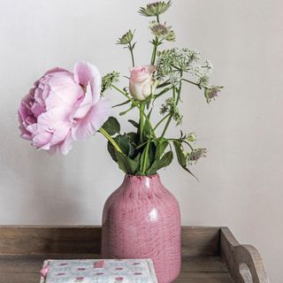 Pale wood tray with pink vase with pink rose and peony beside a blue and pink box. A Grade II listed farmhouse built in 1680 with six bedrooms in Hertfordshire, home of Lauren Richards, founder of a makeup artistry business LC Make Up, and her husband Charlie, three children and two dogs