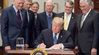 the president of the united states sits at a desk to sign a directive with a moon rock atop a lucite cylinder set beside him. a group of lawmakers stand around the president.