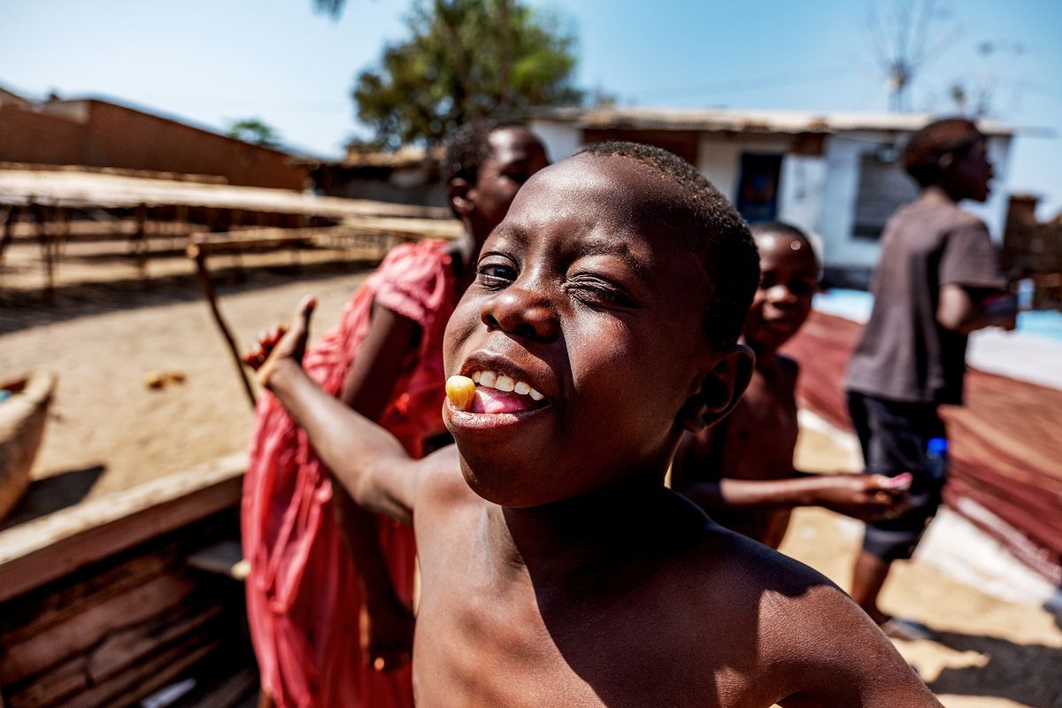Portrait of. a boy having a nut in his mouth, in the background, children play outside in a sandy area, with wooden structures and homes in the background
