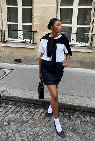 A denim skirt outfit is shown in a photo of a woman on the sidewalk wearing a white t-shirt with a black cardigan, denim mini skirt, white socks, and black ballet flats