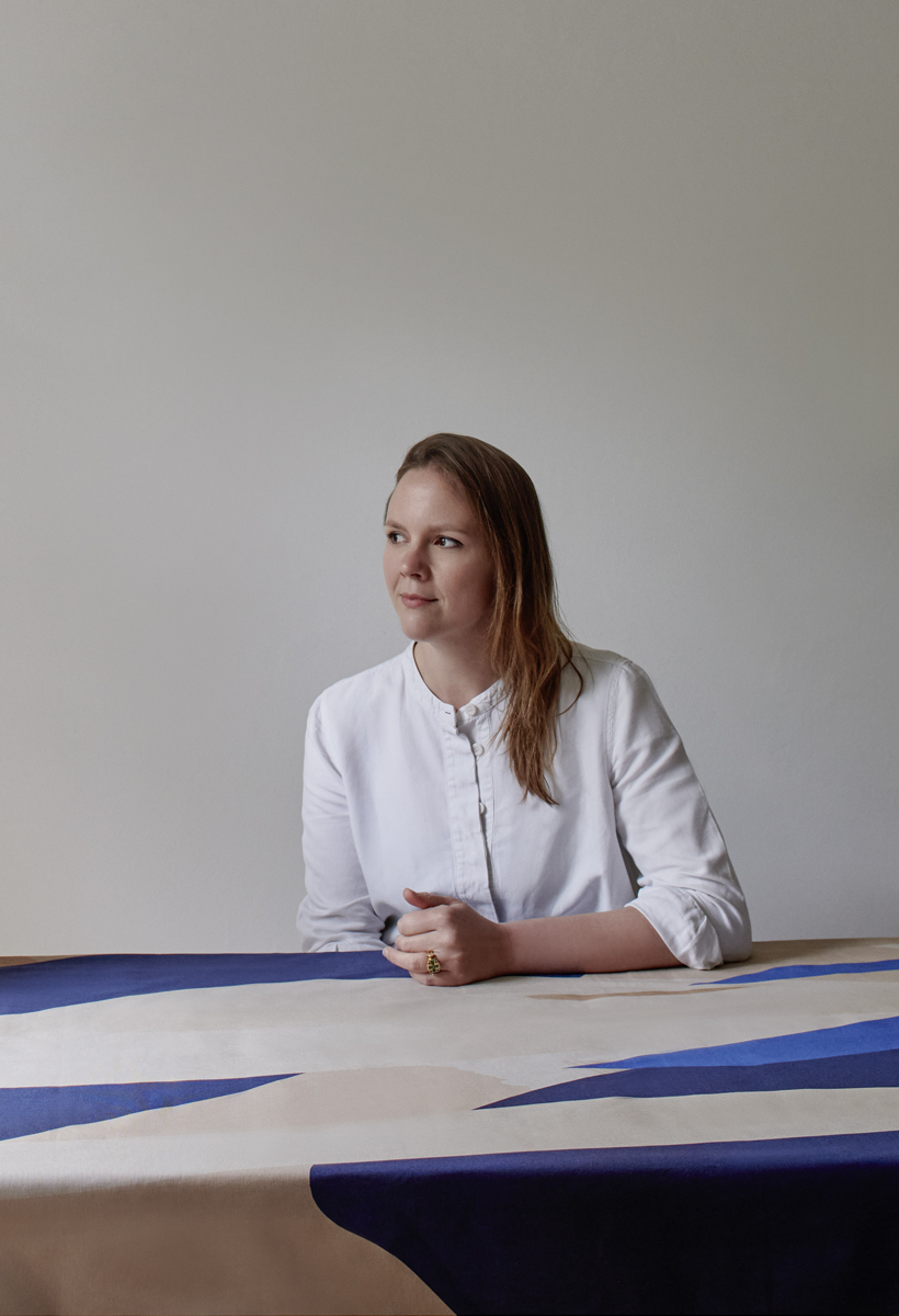 Fiona Ginnett headshot, a woman with auburn hair sitting at a table with a patterned fabric tablecloth on it, resting her arm on the table staring off camera wearing a white shirt