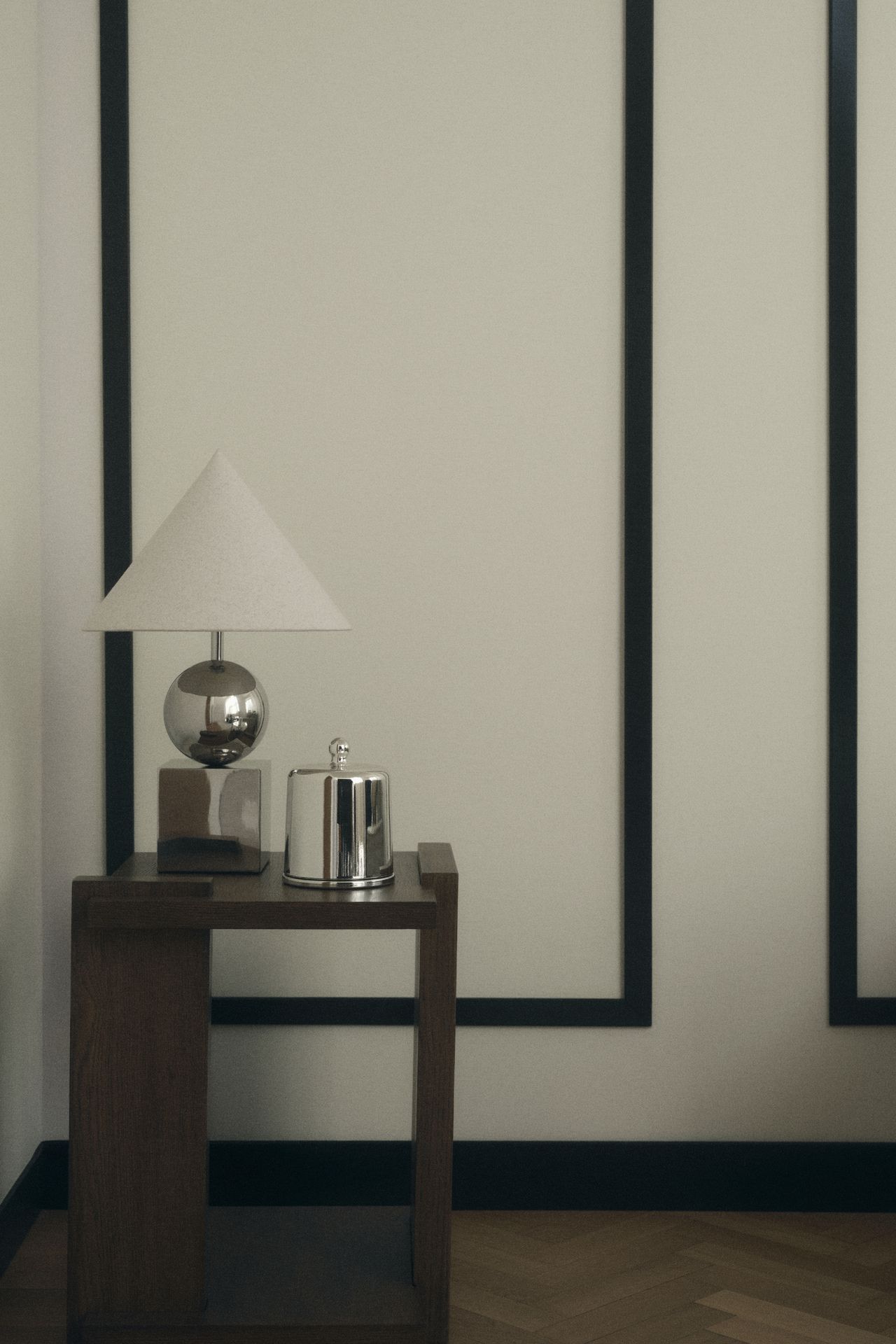 Image of a wooden side table with a silver lamp and silver box on it in front of a white wall with black molding.