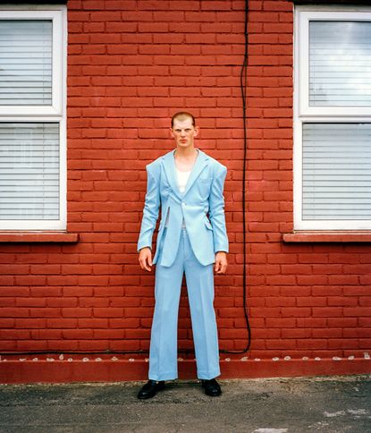 Daytime image of a man in a light blue suit with tie fastening against a red brick wall, white framed windows with white venetian blinds either side