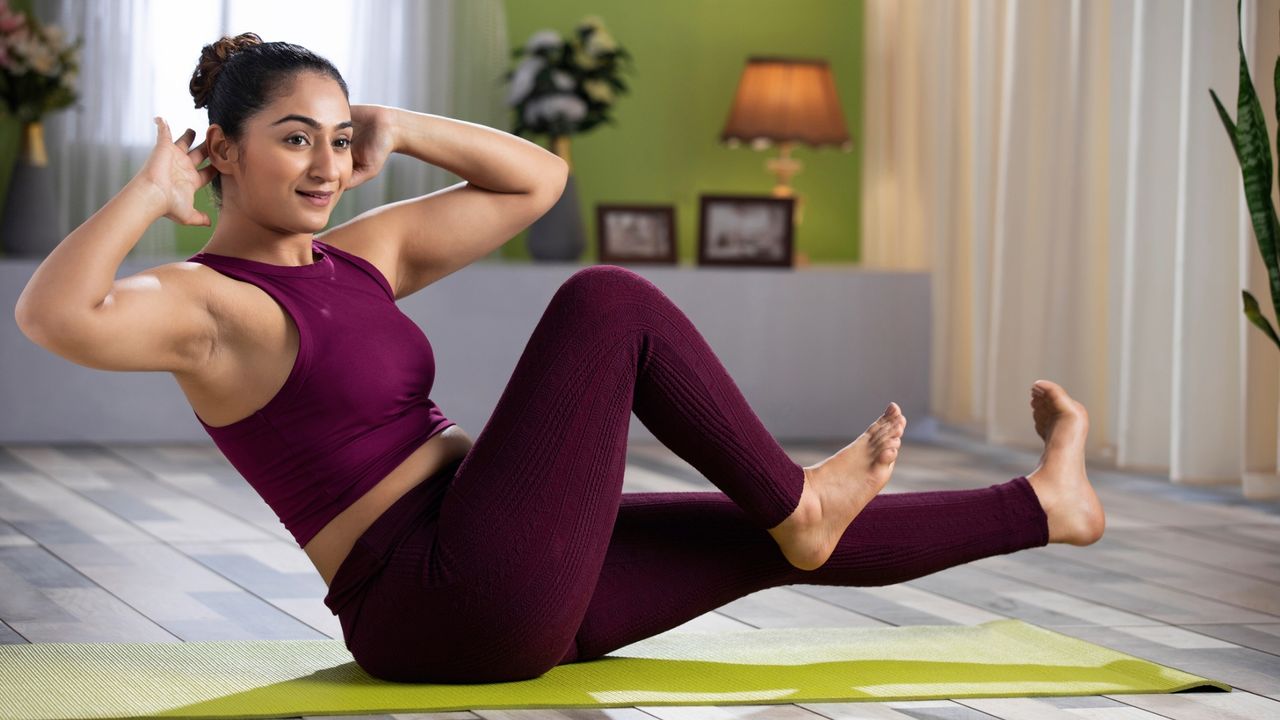 woman wearing wine-coloured leggings and crop top performing a bicycle crunch core exercise head turned towards the camera in a living room setting on a lime green exercise mat. 