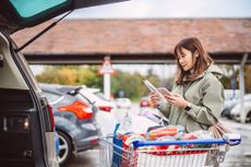 Woman standing beside a shopping cart outside a supermarket, checking her smart phone while reviewing a paper receipt