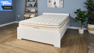 The Saatva Classic mattress photographed on a white bedroom in our blue walled Sleep Lab during the testing process and with a blue Tom's Guide 5 star review rating badge overlaid in the top left hand corner of the image