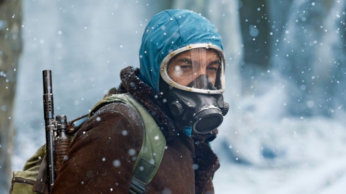 Scene from El Eternauta: A person wearing a gas mask and a blue hood over a brown, fur-lined coat looks directly forward in a snowy, outdoor setting.
