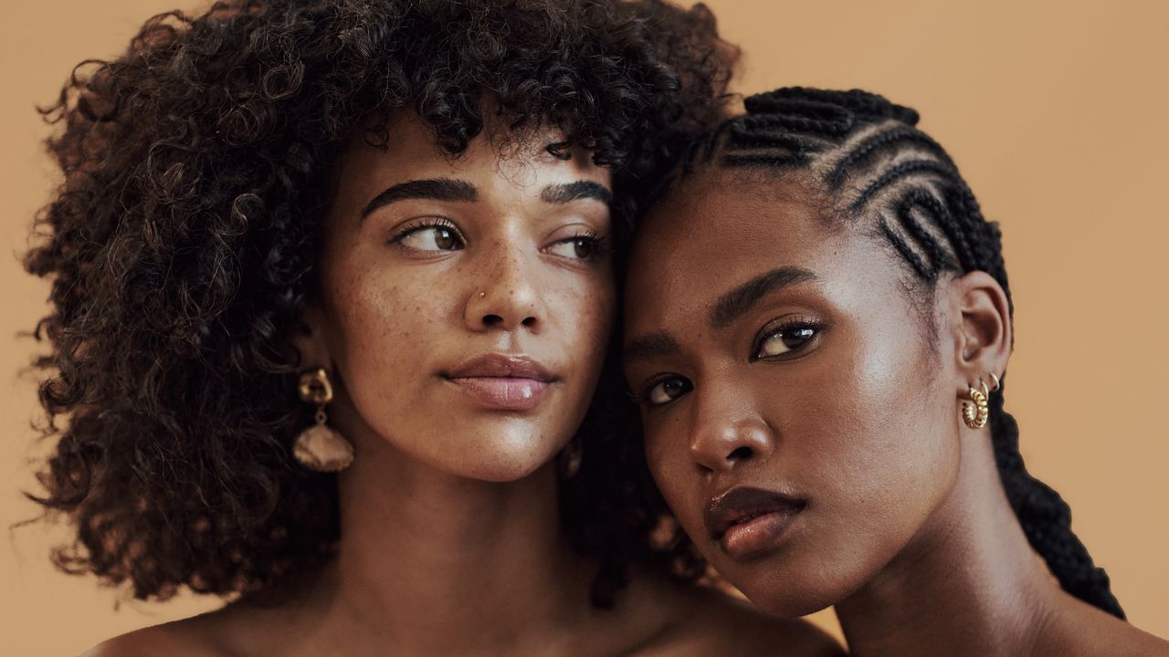 two women with fresh and bright skin leaning together, one with curly hair and the other with braids