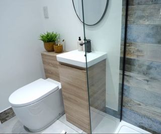 A grey tiled shower wall, white toilet and sink with a wooden cabinet