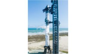 a tall silvery-white rocket stands at a seaside launch pad with sand and the ocean in the background.