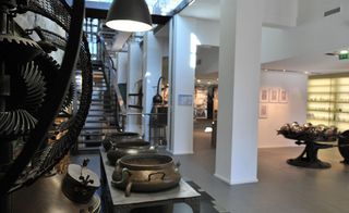 Lobby of the Musée International de la Parfumerie, Grasse, white walls and columns, grey floor, black curved stairwell, display units lit up, black hanging wall light, display items