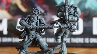 Two unpainted Space Marine models ready their weapons, with the Warhammer 40K Armageddon box set visible behind them