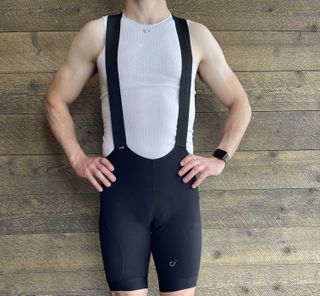 cyclist stood infront of a wood clad wall modelling Velocio Utility bib shorts in black