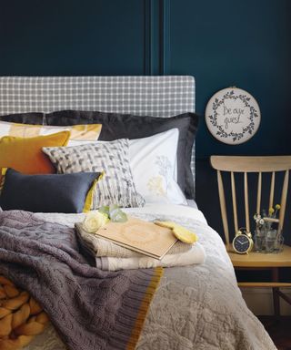 Close up of a guest bed with a variety of throws, cushions and towels. Grey and gold colours with midnight blue walls. Guest Bedroom treats and comforting accessories.