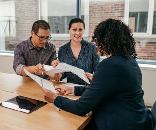 3 people in meeting, 2 women and one man looking at documents in office with wooden meeting table and large glass windows