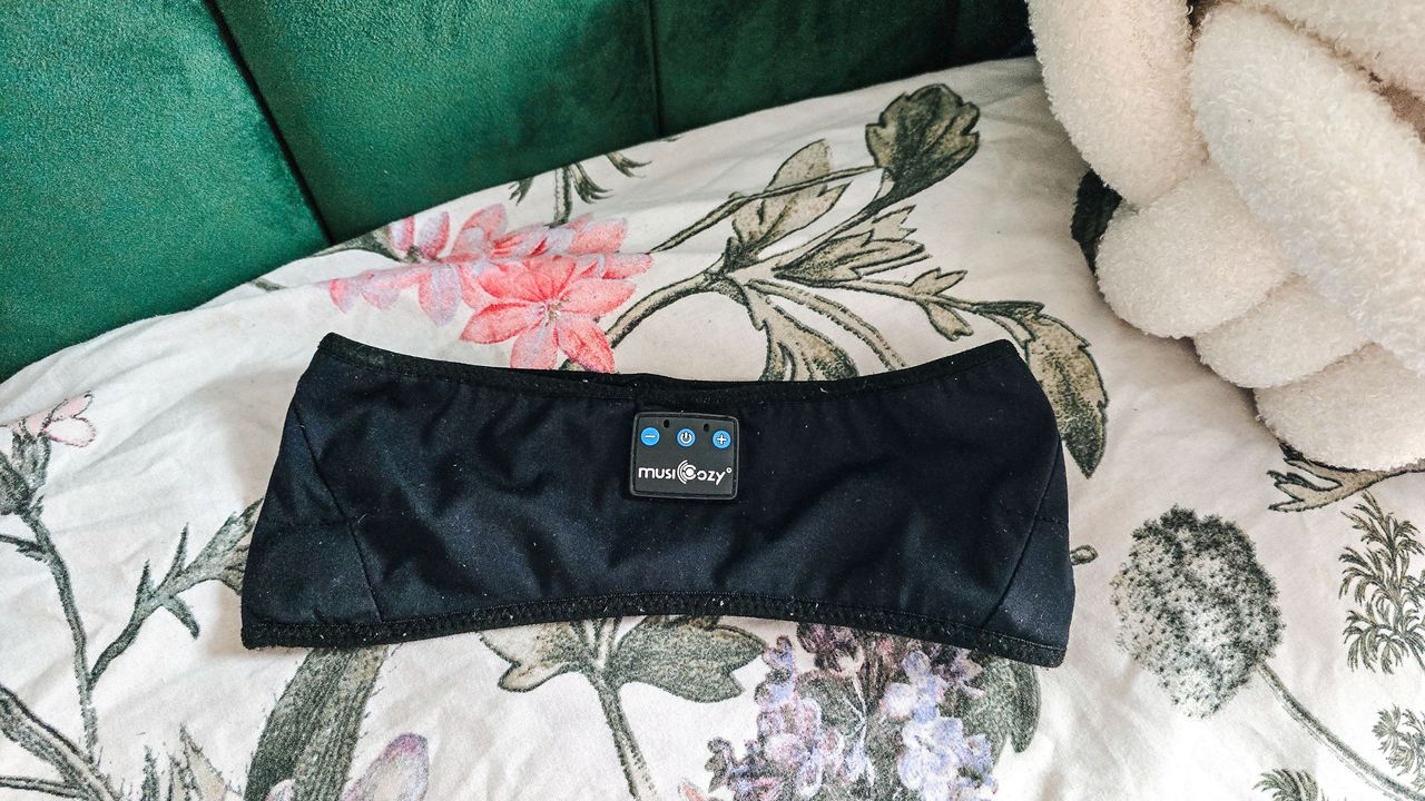 We love sleep mask with headphones. Here is a black rectangular sleep mask lying on top of a white pillow with pink flowers and green stems, with a dark green headboard behind it