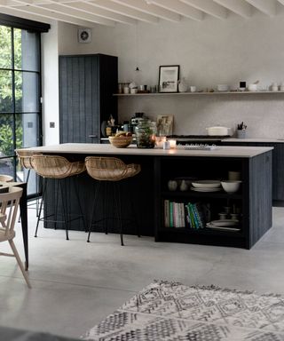 A kitchen with black wooden kitchen island with concrete countertop, cane barstools and Berber carpet