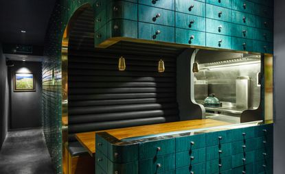 Interior view of Cure featuring dark grey walls with framed wall art and a teal, black and gold pod with wooden dining table and gold pendant lights. There is also a partial view of the kitchen