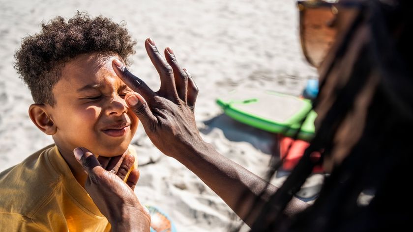 A tan woman wearing sunglasses with long hair applies a dot of sunscreen to a young tan boy who scrunches up his face in response. A beach with toys is blurred but visible in the background. 