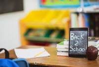 "Back 2 School" written on a digital tablet placed on a brown desk with books, a pencil, paper and an apple (fruit) on the desk as well.