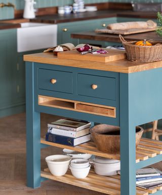 A teal blue kitchen island with wooden shelves underneath with books and white bowls underneath it and a chopping board on top