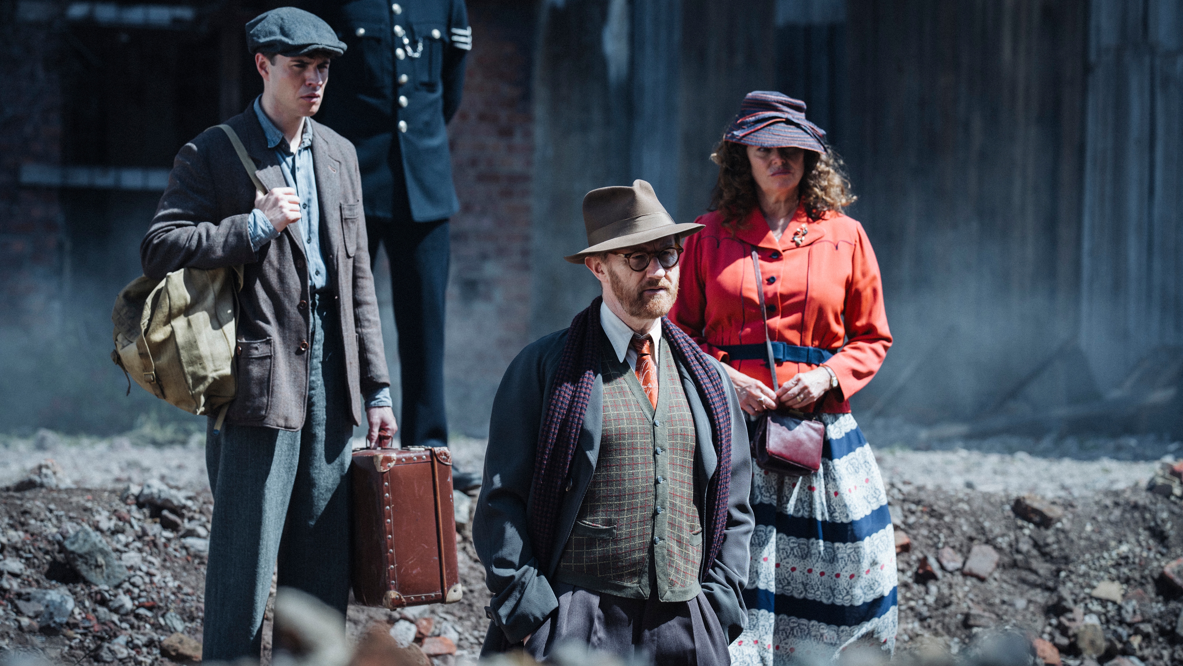 Jack (Connor Finch), Book (Mark Gatiss) and Trottie (Polly Walker) attend a bomb site, standing in a crater in the ground. Jack has a satchel over his shoulder and a small suitcase in his hand, wearing ill-fitting clothes and a flat cap. Book is wearing a suit with a patterned waistcoat, a long coat over the top with a scarf, and a trilby hat. Trottie is wearing a purple hat, a red jacket, a skirt with spiral patterns of white lace-style print interrupted by dark blue horizontal stripes.