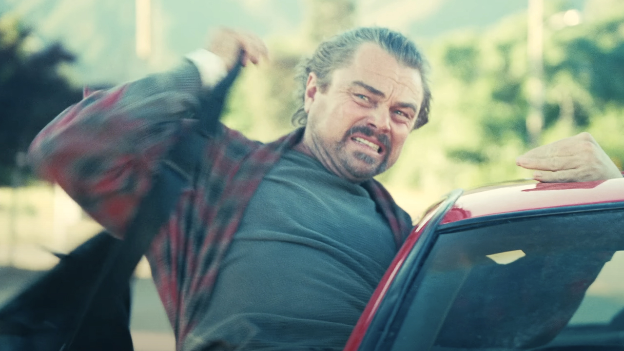 Leonardo DiCaprio dressed in a bathrobe, bag slung over an arm, looks terrified as he hangs out of a moving car in One Battle After Another (2025).