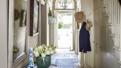 Bright entryway with open front door, white coat stand with coats and hats, white staircase, and side table with green vase of white flowers and white and blue decorative vase.