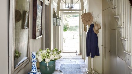 Bright entryway with open front door, white coat stand with coats and hats, white staircase, and side table with green vase of white flowers and white and blue decorative vase.