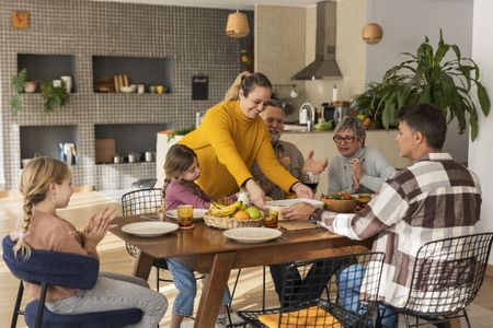 Three generations are living under one roof, or at least enjoying a meal together at home. The adult daughter is serving a dish while her spouse and two children look on. The grandparents are also at the table.
