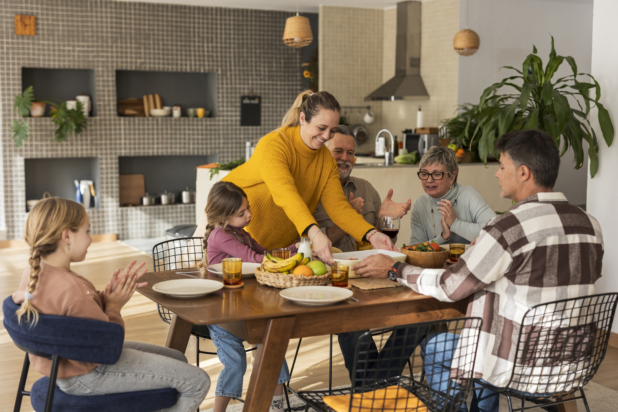 Three generations are living under one roof, or at least enjoying a meal together at home. The adult daughter is serving a dish while her spouse and two children look on. The grandparents are also at the table.