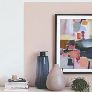 A framed wall print on a painted pink block on the wall with complementing vases next to it