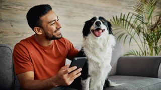 Man smiling at dog he's sat next to on couch while holding phone