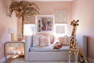 blush pink girl's bedroom with pale gray couch, side table, artwork, blinds, large gold tree floor lamp, toy giraffe, rug,