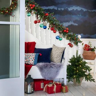 a hallway with blue paint pattern walls, white staircase with christmas garland and baubles along the railing, a white bench seat with red, blue and white cushions, red and gold wrapped presents and a mini christmas tree with fairy lights