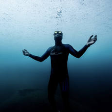 Man in a wetsuit and goggles holding his breath underwater